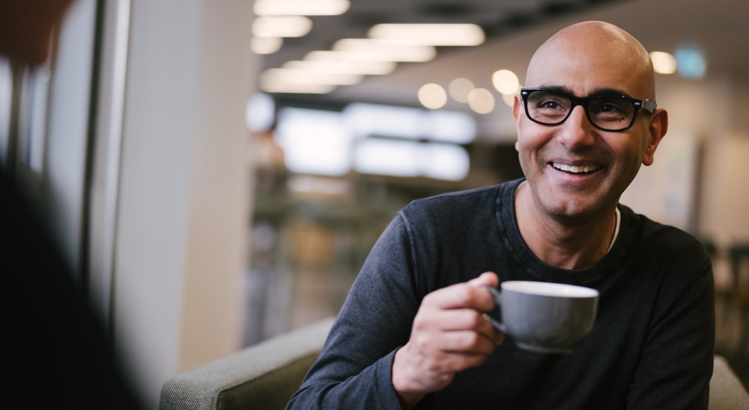 Amjad with a hot drink in the office cafe, smiling at someone just off-screen