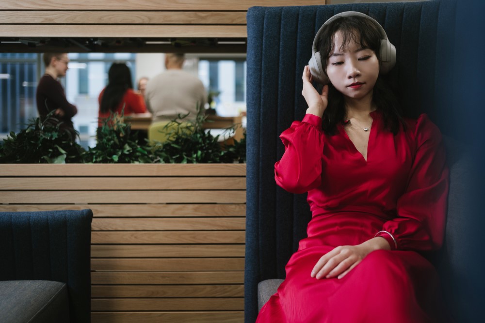 Janet relaxing and listing to music on her headphones in the office with colleagues talking in the background