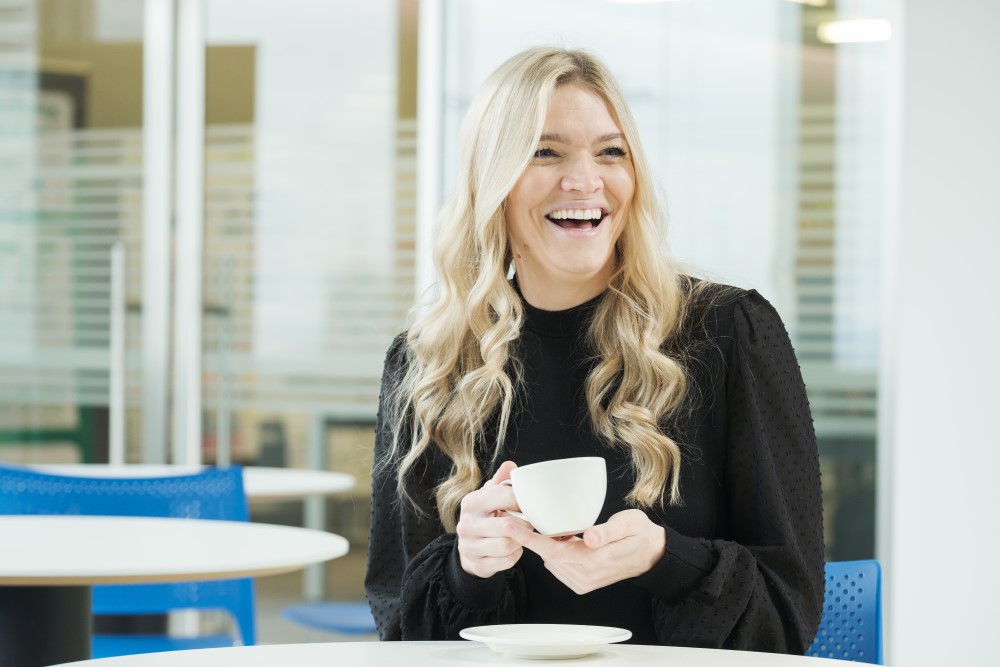 Nicola sitting in an office cafe holding a cup of tea and smiling broadly.