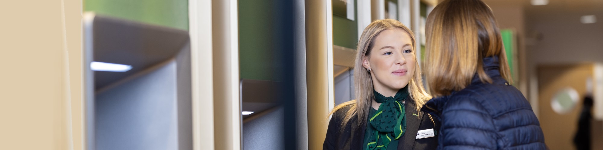 Lloyds Bank collague speaking to a lady in front of a row of cashpoints in a branch.