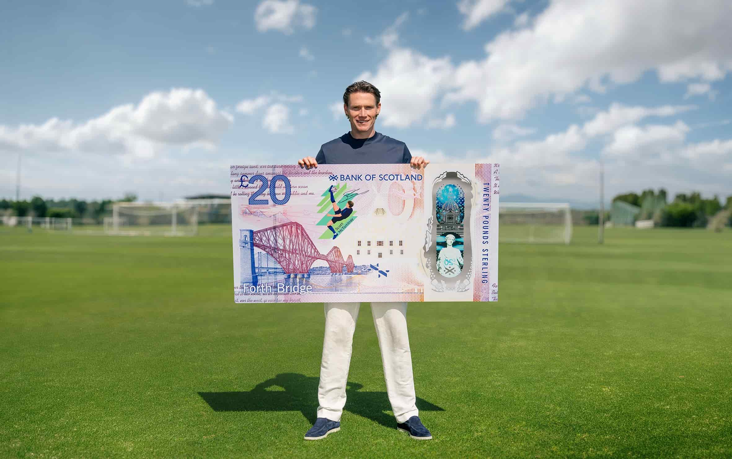 Scott McTominay standing on a football pitch holding an oversized Bank of Scotland £20 note featuring a football-themed design, with goalposts and a clear blue sky in the background.
