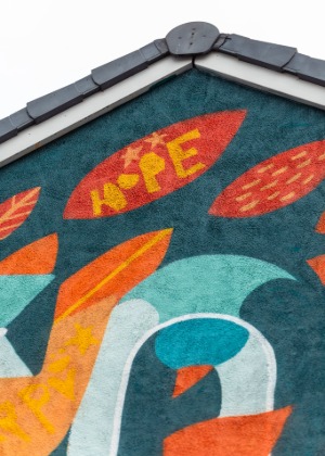 A close-up of the top corner of the mural on a building wall. The background is painted dark teal with several bright orange and red leaf shapes. One large red leaf near the roof edge has the word “HOPE” painted in yellow with small decorative stars. The roof tiles and trim of the building are visible at the top of the image.