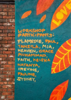 A section of the mural showing a brick wall on the left and turquoise-painted wall on the right. In orange hand-painted text, the words “Workshop Participants:” are followed by a list of names: Plamedie, Paul, Tanzila, Mia, Heaven, Grace, Muhammad, Faith, Keisha, Natanya, Irevine, Pauline, and Sydney. Bright orange and yellow leaf shapes decorate the background.