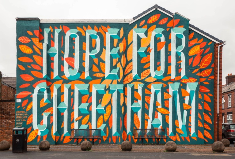 A large, brightly coloured mural painted across the side of a brick building, featuring the words “HOPE FOR CHEETHAM” in bold, light-blue lettering. The background is filled with orange, red and teal leaf shapes, with smaller words like “hope,” “grow,” and “love” written inside some leaves. Houses and rooftops are visible behind the building.