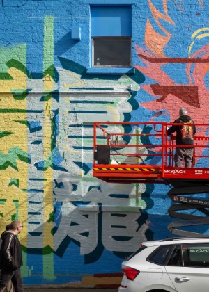 Artist painting large Chinese characters on a blue wall from a cherry picker while a passer-by walks past on Grenville Street South, Liverpool.